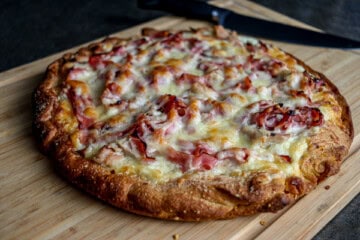 A chicken cordon bleu pizza with cheese and bacon on a wooden cutting board