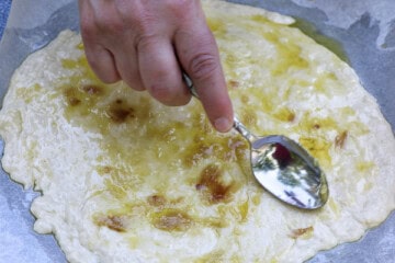 A person is using a spoon to spread olive oil/garlic on to the dough